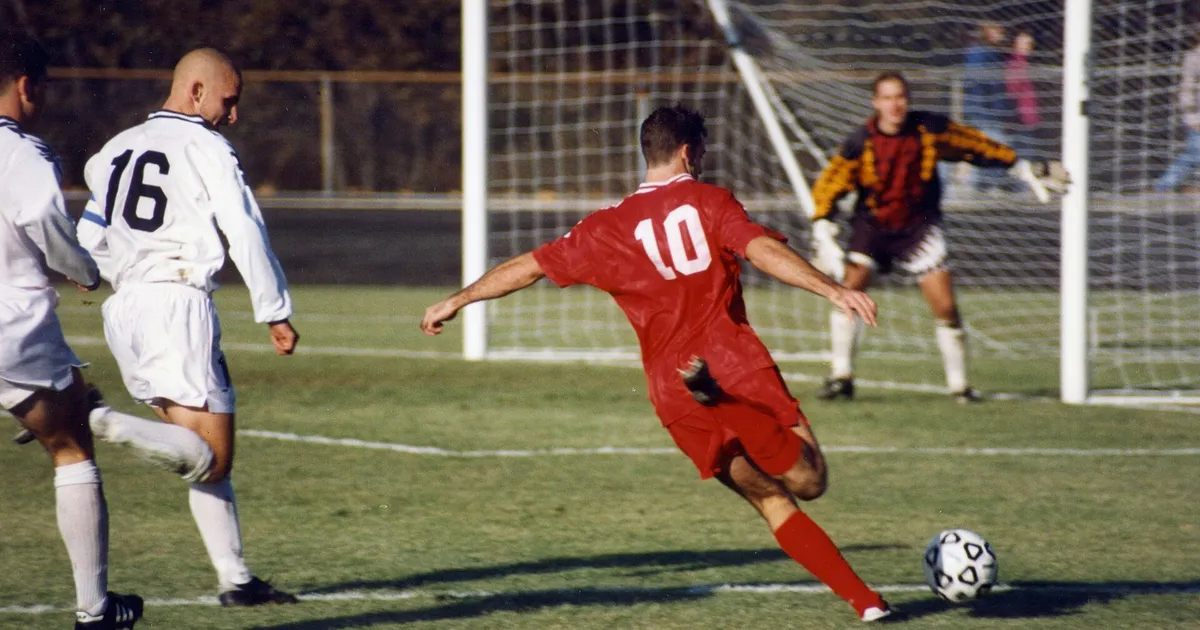 Association football (soccer) , Bloomington, Indiana. The offensive player (in red) is Chris Klein of the Indiana Hoosiers. He has sprinted past two defenders (in white) and is about to shoot the ball at the goal or pass it across the field to a teammate in front of the goal.