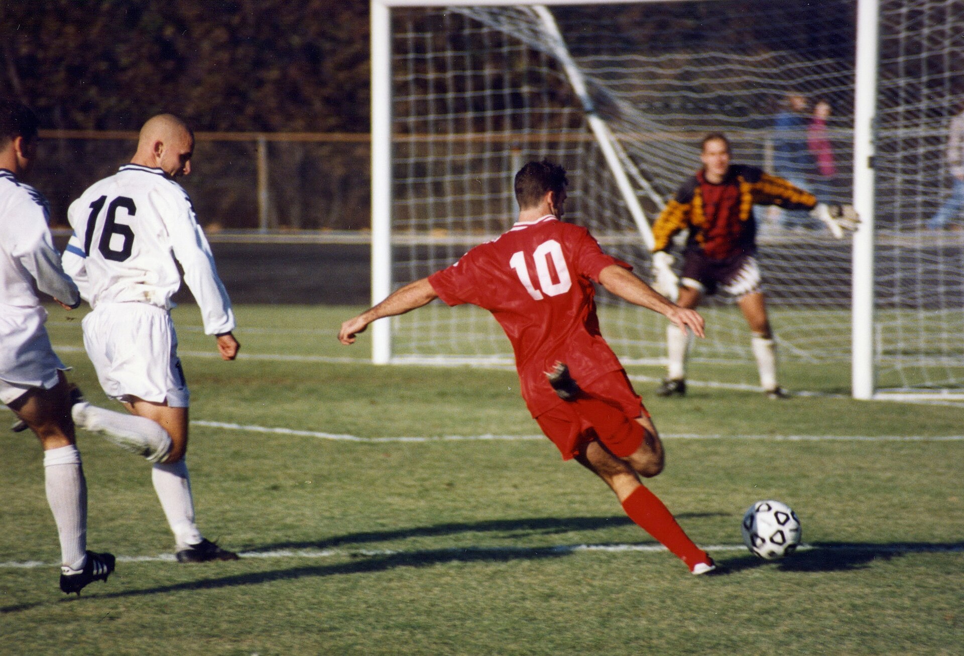 Association football (soccer) , Bloomington, Indiana. The offensive player (in red) is Chris Klein of the Indiana Hoosiers. He has sprinted past two defenders (in white) and is about to shoot the ball at the goal or pass it across the field to a teammate in front of the goal.